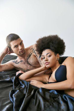 Téléchargez les photos : Bel homme regardant la caméra et couché dans le lit avec sa belle petite amie afro-américaine - en image libre de droit