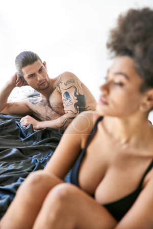 Foto de Centrarse en el hombre guapo con tatuajes acostado en la cama junto a su novia afroamericana borrosa - Imagen libre de derechos
