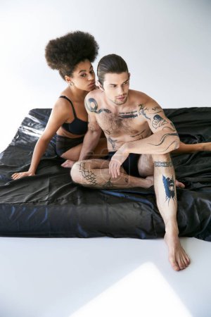 Téléchargez les photos : Bel homme avec des tatouages assis sur le lit à côté de sa petite amie afro-américaine, couple sexy - en image libre de droit