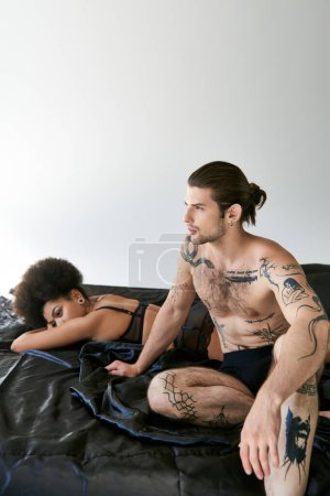 Téléchargez les photos : Jeune femme afro-américaine couchée au lit avec son petit ami assis à côté d'elle, couple sexy - en image libre de droit