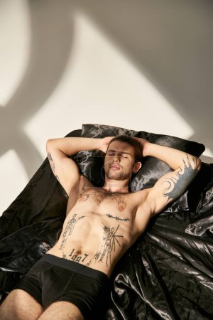 young tattooed male model with ponytail lying in bed with hands behind head with closed eyes