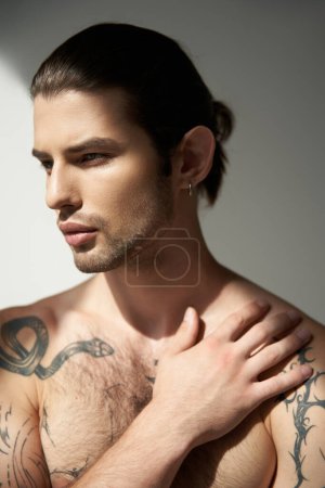appealing young man with tattoos and ponytail posing with his hand on chest and looking away