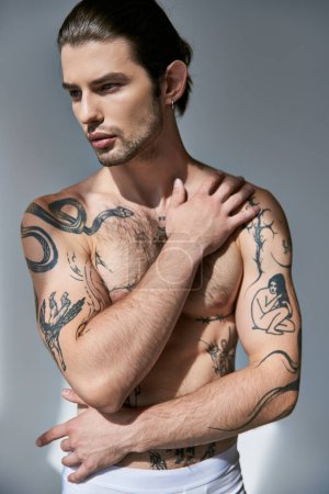 Foto de Joven guapo con tatuajes y cola de caballo posando con la mano en el pecho y mirando hacia otro lado - Imagen libre de derechos