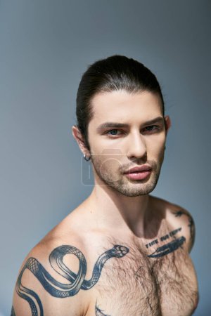 Foto de Guapo joven modelo masculino con tatuajes y cola de caballo mirando a la cámara y posando sobre fondo gris - Imagen libre de derechos