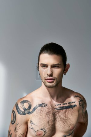 good looking tattooed man with ponytail and earring posing on gray backdrop and looking away