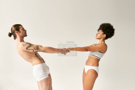 junges multirassisches sexy Paar in Unterwäsche, Händchen haltend und einander sinnlich betrachtend