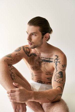 Foto de Hombre joven y guapo tatuado con cola de caballo en ropa interior sentado en el suelo y mirando hacia otro lado - Imagen libre de derechos