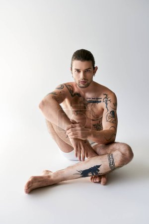 Foto de Buen aspecto joven tatuado modelo masculino en ropa interior sentado en el suelo y mirando a la cámara - Imagen libre de derechos