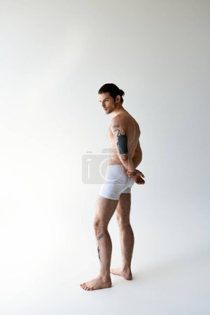 sexy starker Mann mit Pferdeschwanz und coolen Tattoos posiert in bequemer Unterwäsche auf Ecru-Hintergrund