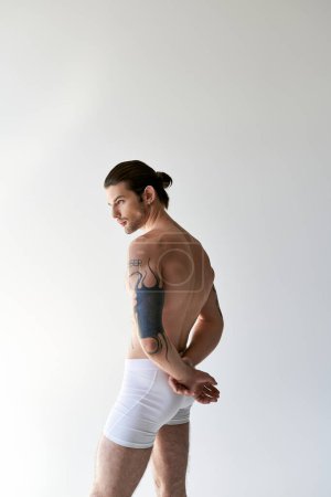 junger starker Mann mit Pferdeschwanz und coolen Tattoos in bequemer Unterwäsche posiert auf Ecru-Hintergrund