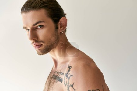 verführerischer junger Mann mit Pferdeschwanz und coolen Tattoos in bequemer Unterwäsche posiert auf Ecru-Hintergrund