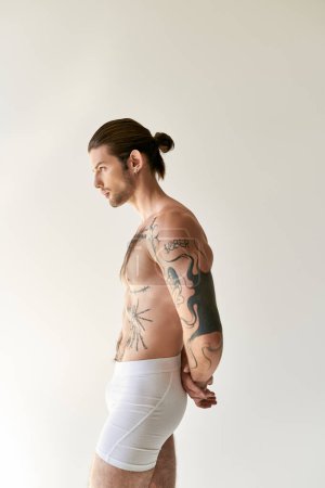 Foto de Joven hombre guapo con cola de caballo y tatuajes frescos en ropa interior posando sobre fondo crudo - Imagen libre de derechos