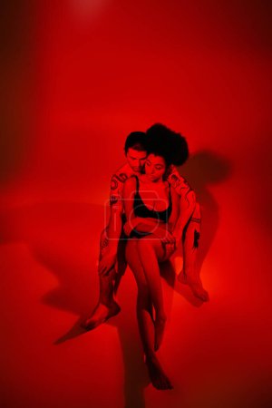Téléchargez les photos : Bel homme avec des tatouages posant avec sa petite amie afro-américaine dans les lumières rouges, couple sexy - en image libre de droit