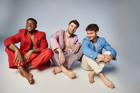 fröhliche multiethnische Männer in hellen Anzügen, die mit überkreuzten Beinen auf dem Boden sitzen und freudig lächeln