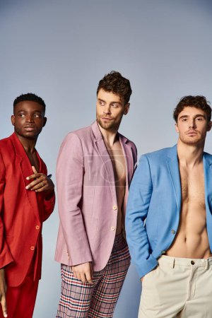 attraktive multirassische Freunde in farbenfrohen stilvollen Anzügen posieren zusammen auf grauem Hintergrund, Mode