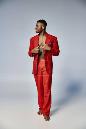 hombre afroamericano guapo en traje rojo elegante posando sobre fondo gris, concepto de moda