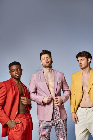 tres diversos hombres en vibrantes trajes desabotonados posando juntos sobre fondo gris, concepto de moda