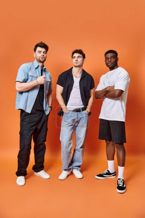 three appealing multicultural friends in casual urban outfits posing together on orange backdrop