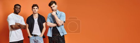 handsome multicultural friends in casual urban outfits posing together on orange backdrop, banner