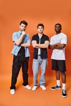 three handsome multiracial friends in casual urban outfits posing together on orange backdrop