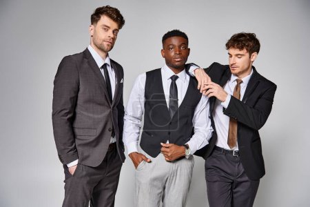 attraktive junge multikulturelle Freunde in schicken Business-Anzügen posieren gemeinsam vor grauem Hintergrund