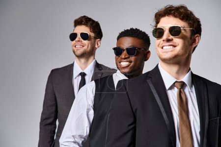 Téléchargez les photos : Joyeux divers amis en tenue décontractée d'affaires avec des lunettes de soleil posant ensemble sur fond gris - en image libre de droit