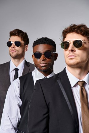 Téléchargez les photos : Beaux modèles masculins multiraciaux dans des costumes élégants intelligents avec des lunettes de soleil posant sur fond gris - en image libre de droit