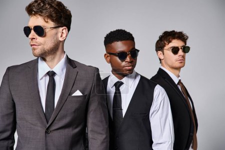 Téléchargez les photos : Beaux modèles masculins multiraciaux dans des costumes élégants intelligents avec des lunettes de soleil posant sur fond gris - en image libre de droit