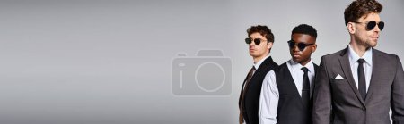 Téléchargez les photos : Bel homme diversifié dans des costumes élégants intelligents avec des lunettes de soleil posant sur fond gris, bannière - en image libre de droit