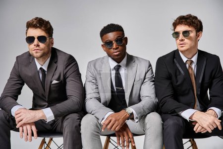 good looking diverse men with sunglasses in business attires sitting on chairs on gray backdrop