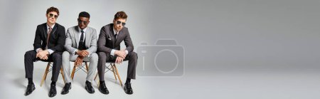 appealing diverse men with sunglasses in business attires sitting on chairs on gray backdrop, banner
