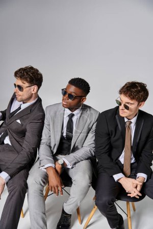 beaux hommes multiraciaux avec des lunettes de soleil en tenue d'affaires assis sur des chaises sur fond gris
