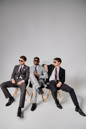 attractive interracial men with sunglasses in smart attires sitting on chairs on gray backdrop