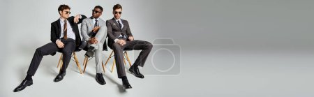 Téléchargez les photos : Beaux hommes interracial avec des lunettes de soleil en vêtements intelligents assis sur des chaises sur fond gris, bannière - en image libre de droit