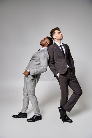 dos atractivos modelos masculinos multiculturales con estilo en trajes inteligentes de negocios posando sobre fondo gris