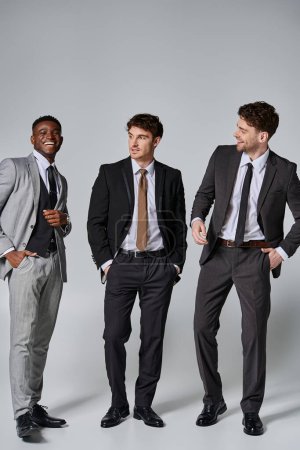 modelos masculinos multirraciales alegres de buen aspecto en trajes elegantes sonriendo sinceramente sobre fondo gris