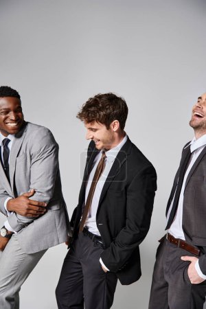 atractivas modelos masculinos multiculturales alegres en trajes inteligentes sonriendo sinceramente sobre fondo gris