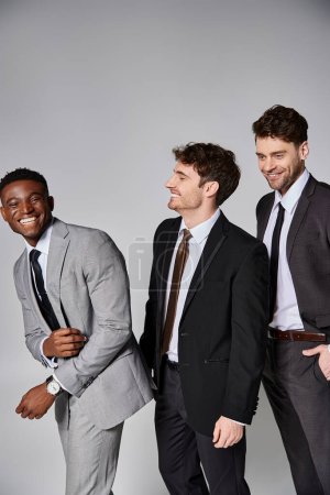 good looking cheerful multicultural male models in smart suits smiling sincerely on gray backdrop