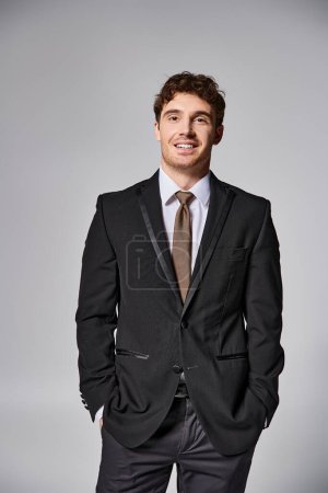 cheerful young man in smart business suit posing on gray backdrop and smiling joyfully at camera