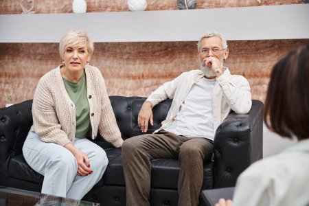 pareja de mediana edad sentado en el sofá de cuero y mirando al psicólogo durante la consulta