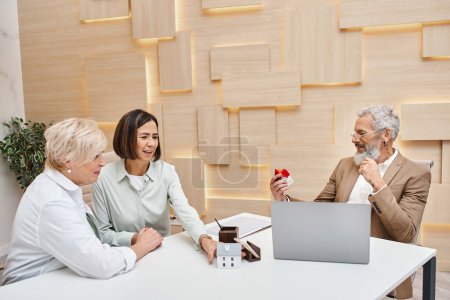 middle aged bearded realtor holding house model near interracial lgbt couple in real estate office