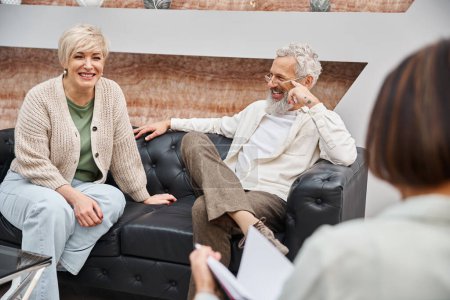 glückliches Paar mittleren Alters auf Ledercouch sitzend und während der Beratung zum Psychologen schauend