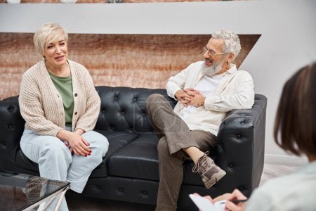 happy middle aged couple sitting on leather and smiling with psychologist during consultation