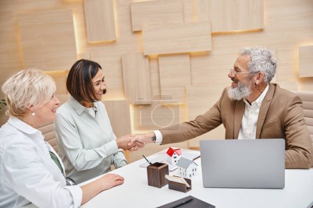 realtor shaking hand with middle aged multiracial woman near lesbian partner  in real estate office