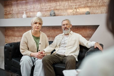 Téléchargez les photos : Attentif couple d'âge moyen assis sur le canapé en cuir et en regardant psychologue pendant la consultation - en image libre de droit
