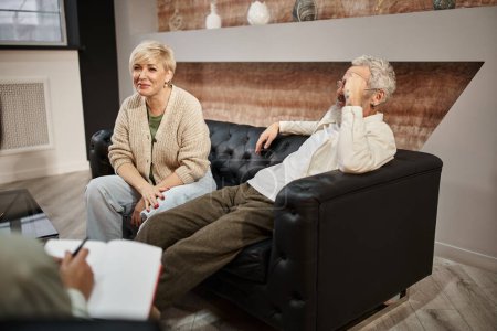 cheerful middle aged woman sitting on leather couch near husband during family therapy session