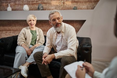 Emotionales Paar mittleren Alters sitzt auf der Couch und spricht während einer Therapiesitzung mit einem Psychologen