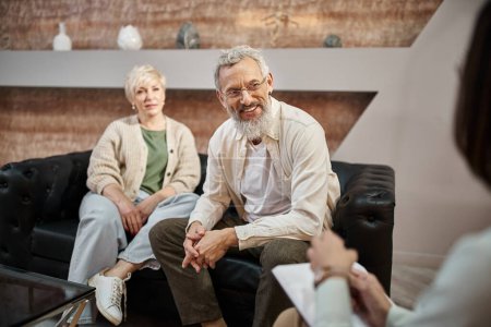 cheerful bearded man sitting on couch and looking at family consultant near wife during session
