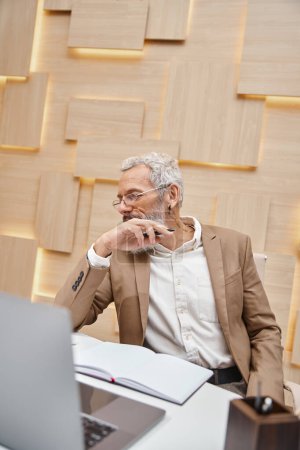 Makler mittleren Alters mit Brille am Laptop im modernen Büro, Stift in der Nähe von Notizbuch