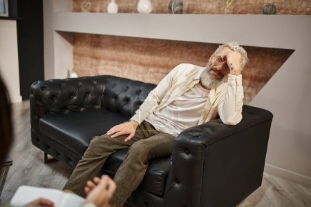 bearded middled aged man sitting on leather couch and looking at psychologist during session